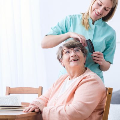 Eine Frau bürstet das Haar einer älteren Dame, umgeben von einer freundlichen und häuslichen Atmosphäre.